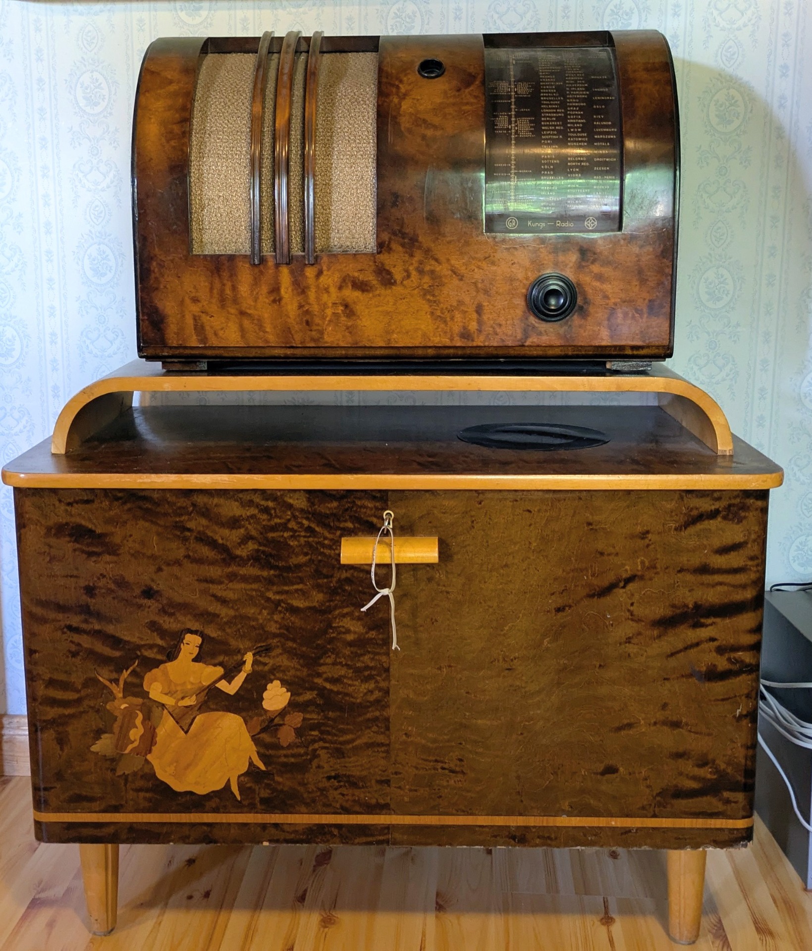 Brun vintage radioapparat med högtalargaller, textpanel och dekorativt motiv på låda, på träbord.